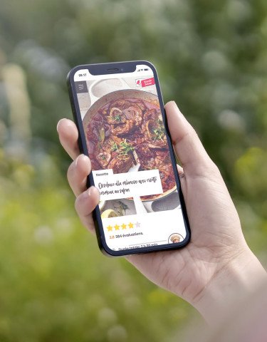 Une main tenant un smartphone à l'extérieur avec une vue floue sur la nature en arrière-plan. L'écran affiche une recette de Viande Suisse pour un « Ossobuco alla milanese », incluant une photo du plat et une évaluation par étoiles.